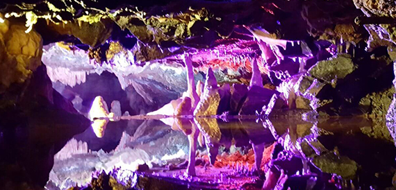 Cave formations lit in purple and pink, reflected in the water below