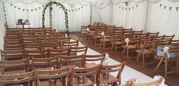 Wedding ceremony aisle in a marquee with a floral arch and wooden chairs.