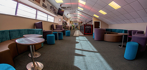 Lounge corridor with teal stools and armchairs beside tall windows.