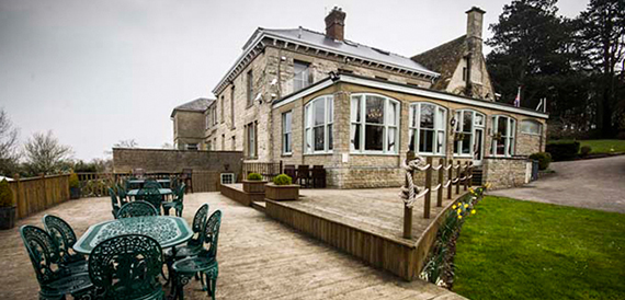 Stone building exterior with a wooden deck terrace and green metal furniture.