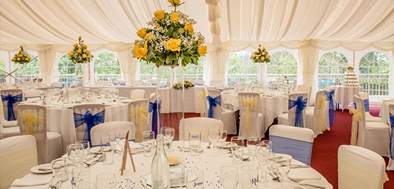 Marquee reception with round tables, blue chair sashes and tall yellow flower displays.