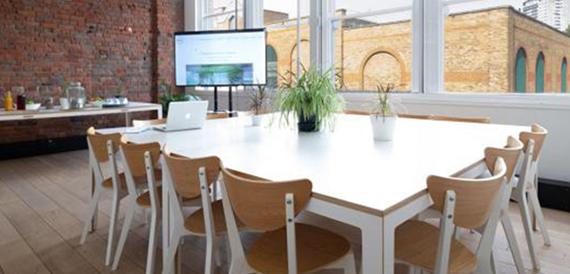 Meeting room with a square white table, wooden chairs and a wall-mounted screen
