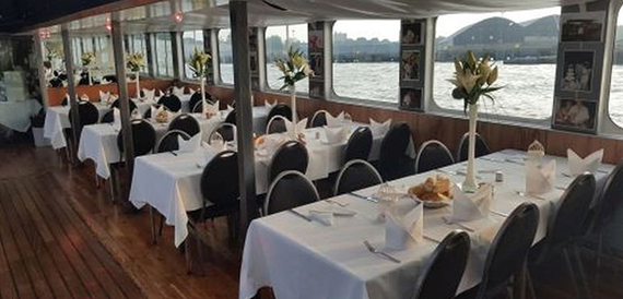 Long dining tables set with white linen along the boat's windowed deck.