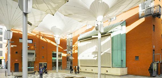 Sunlit courtyard beneath open canopy sails with a few people passing through.