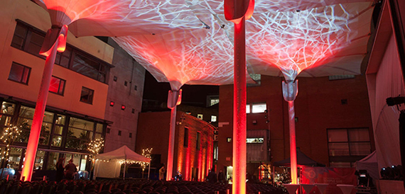 Illuminated tensile canopy structure over an empty courtyard lit red at night.