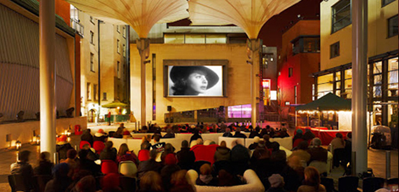 Crowd watching an outdoor cinema screening against a floodlit building facade at dusk.