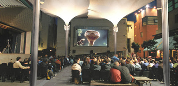 Evening crowd watching a large outdoor screen beneath the canopy roof.