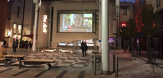 Night view of the square with a cinema screen, The Ark building and benches.