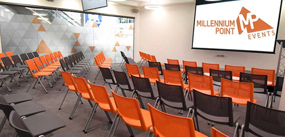 Meeting room set theatre-style with rows of orange and black chairs facing a screen.