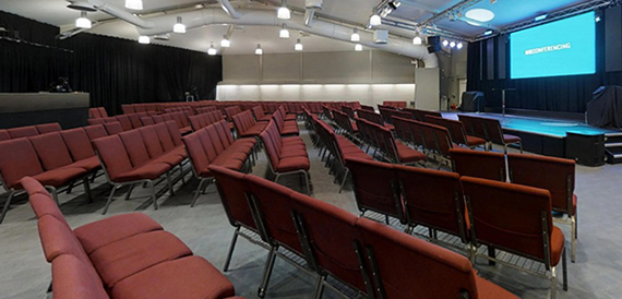 Auditorium set theatre-style with red seating facing a stage and screen.