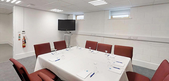 Meeting room with a boardroom-style table, red chairs and a wall-mounted screen.