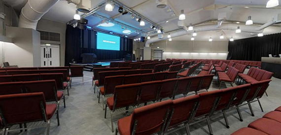 Auditorium set theatre-style with red seating and a lit stage screen ahead.