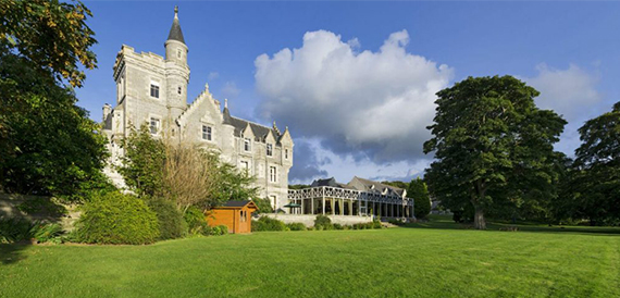 Exterior of the turreted stone mansion house with lawns and mature trees.