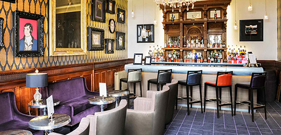 Hotel bar with a dark wood counter, purple sofas and a gallery of framed portraits.