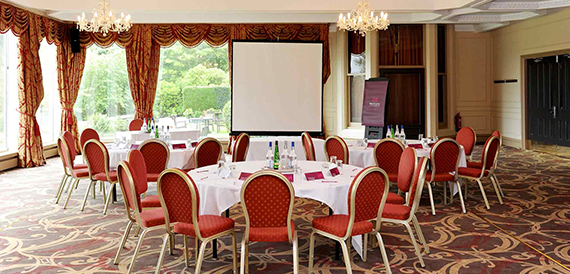 Meeting room laid out cabaret-style with red chairs beneath chandeliers and a screen.