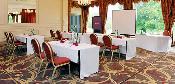 Meeting room with u-shaped tables, red chairs and tall curtained windows.