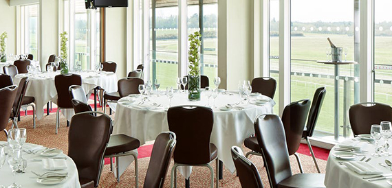 Round dining tables with white linen set beside floor-to-ceiling windows over the racetrack
