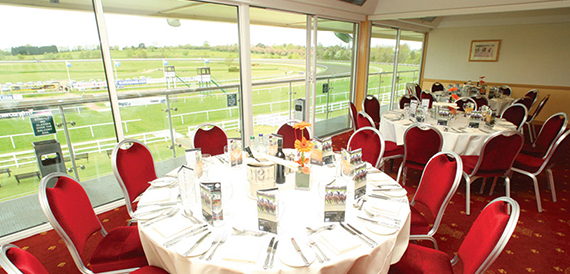 Racecourse-view dining room with round tables, red chairs and race day programme centrepieces