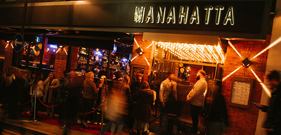 Exterior entrance at night with a queue of guests beneath illuminated Manahatta signage