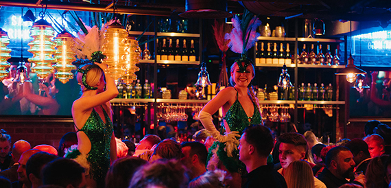 Two performers in green sequinned outfits with feather headdresses among the crowd