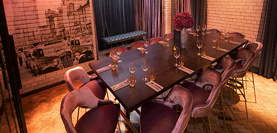 Private dining table with pink velvet chairs set with glassware beneath a brick wall mural