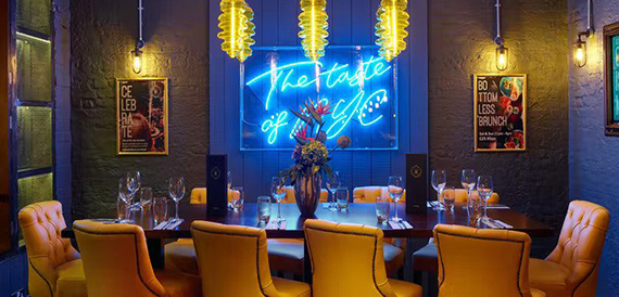 Private dining table with mustard chairs beneath a neon sign and framed artwork