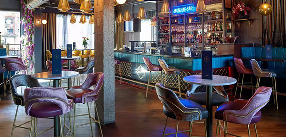 Bar area with pink and purple velvet chairs around round tables beneath a neon Highline Bar sign