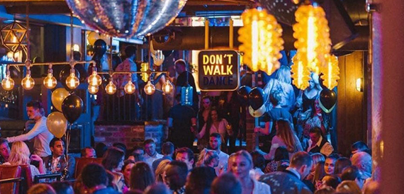 Busy bar scene with guests dancing beneath a neon Don't Walk Dance sign and balloons