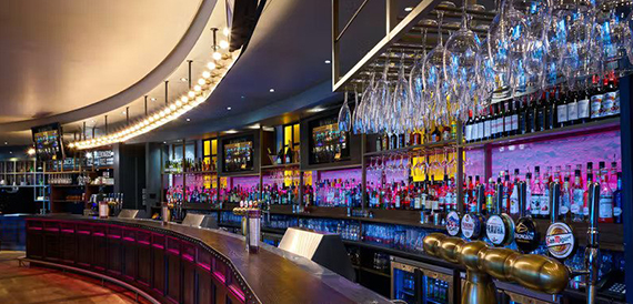 Curved bar counter with hanging wine glasses above a pink-lit bottle display