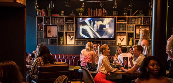 Guests socialising at a booth beneath a wall-mounted screen and shelving display