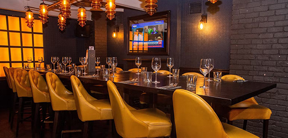 Private dining table with mustard chairs beneath amber pendant lights and a wall screen