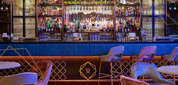 Bar counter with hanging glassware above blue-lit banquette seating and an M monogram