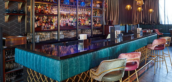 Teal bar counter with pink and grey velvet stools in front of illuminated shelving