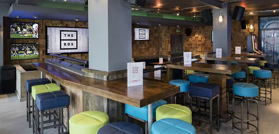 Bar counter with colourful stools beneath screens reading 'The Box'
