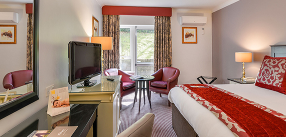 Hotel bedroom with a red throw, burgundy armchairs and a wall-mounted television.