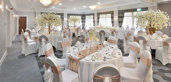 Wedding banqueting room with white chair covers, rose gold sashes and blossom tree centrepieces.