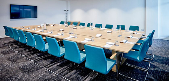 Boardroom with a long table, teal chairs and a wall-mounted screen.