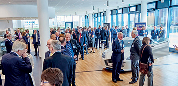 Delegates networking beside a car display in a bright atrium event space.