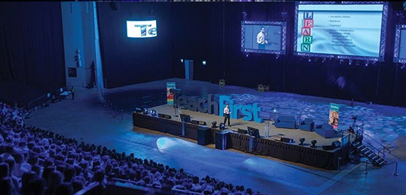 Speaker on stage addressing a seated audience beneath large screens and blue lighting.