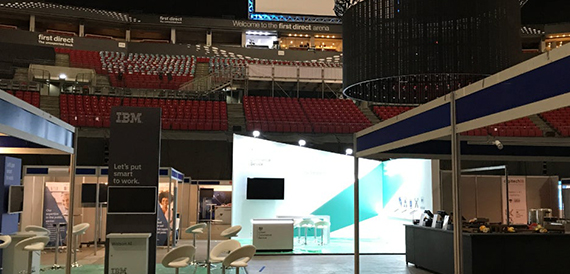 Empty arena seating overlooking exhibition stands and a first direct branded wall.