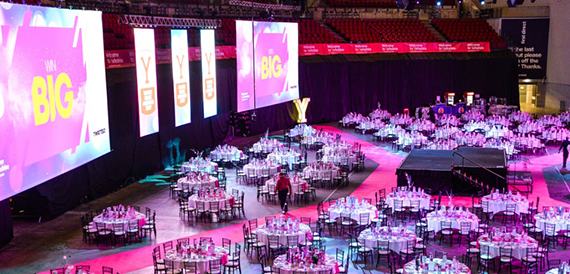 Arena floor set with round banquet tables beneath large pink event screens.
