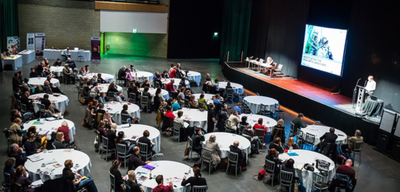 Conference audience seated at round tables facing a stage with speakers