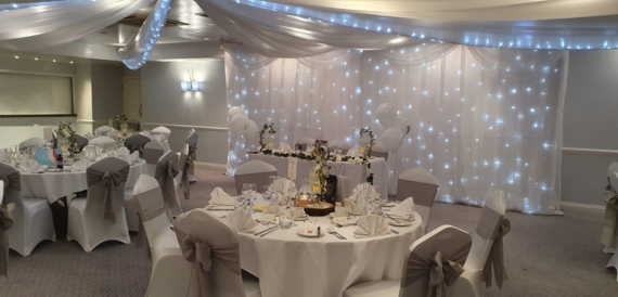 Wedding reception room with grey chair covers, sashes and a fairy-light backdrop.