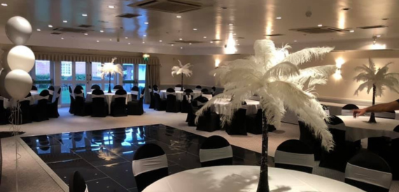 Function room set for a party with black chair covers, ostrich feather centrepieces and balloons.