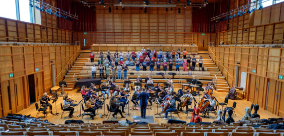 Wood-panelled concert hall with an orchestra and choir rehearsing on stage
