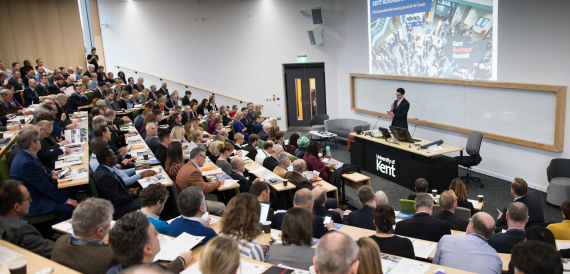Full lecture theatre with a speaker presenting to a seated audience