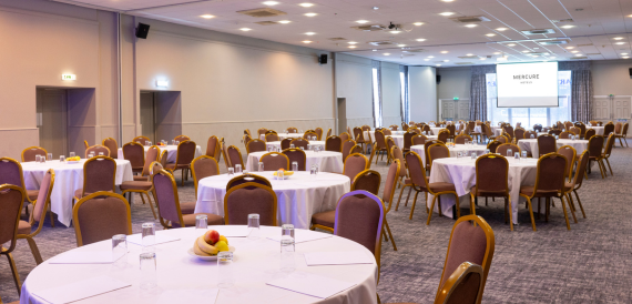 Large banqueting room set for a conference with round tables and a screen.