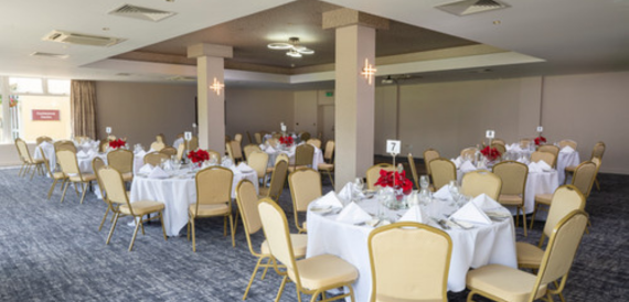 Banqueting room set for a dinner with round tables and red floral centrepieces.