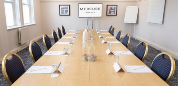 Boardroom with a long table, gold-trimmed navy chairs and a wall-mounted screen.