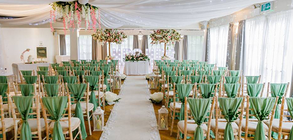 Wedding ceremony aisle with sage green chair sashes and floral displays overhead.